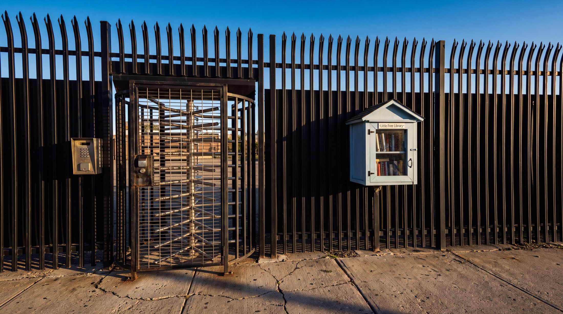 A tall black steel anti-climb security fence with curved spear tops, photographed in low golden light. A full-height interleaved steel exit turnstile sits caged in the fence on the left, with an intercom keypad beside it. Mounted further along the fence, small against it, is a white Little Free Library box with a glass door and two shelves of books. Cracked concrete underfoot, weeds in the seams, deep blue sky above.